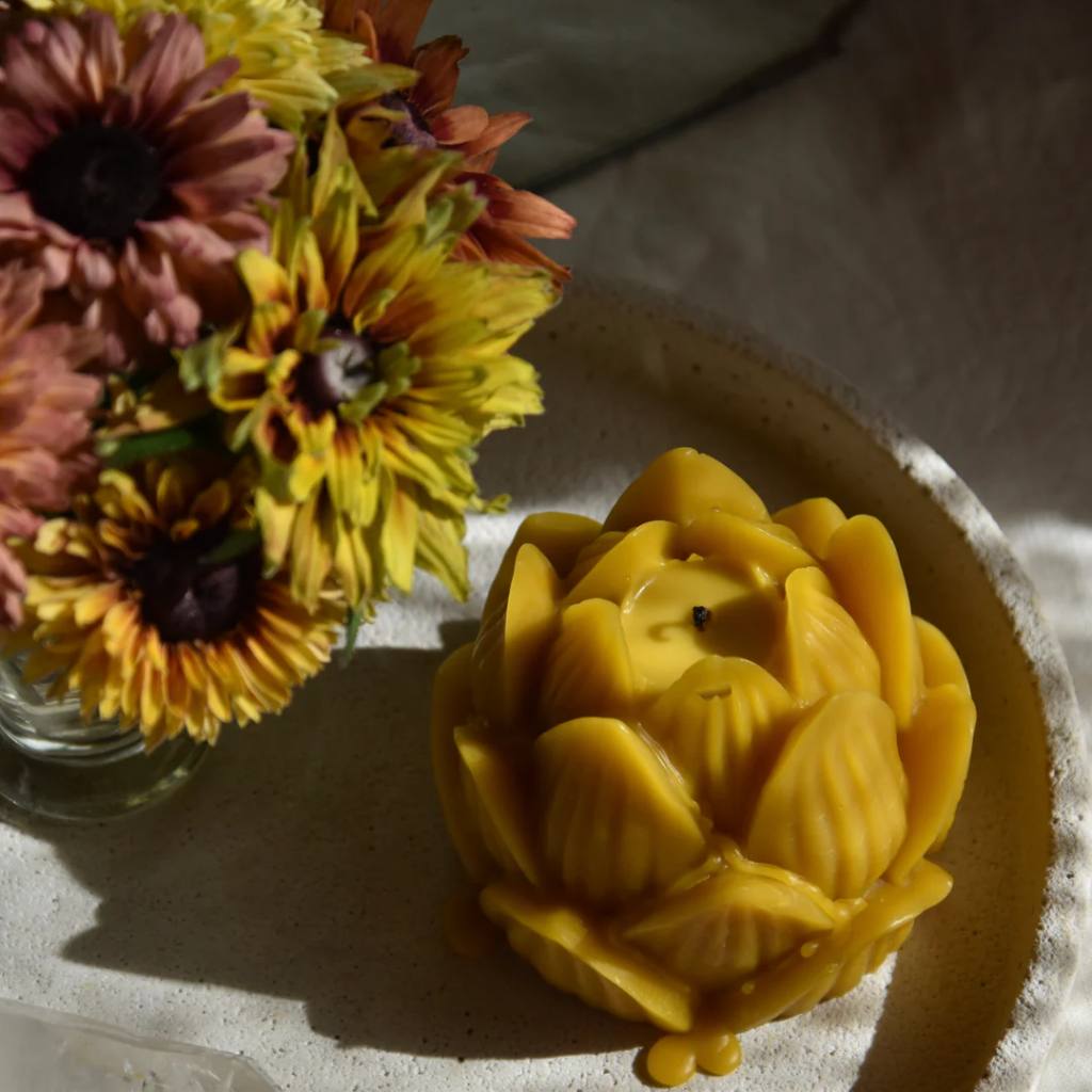 A large, natural beeswax candle shaped like a lotus flower, lit and glowing warmly against a dark background. The intricate petal details of the lotus are highlighted by the soft, ambient light, creating a serene and calming atmosphere.