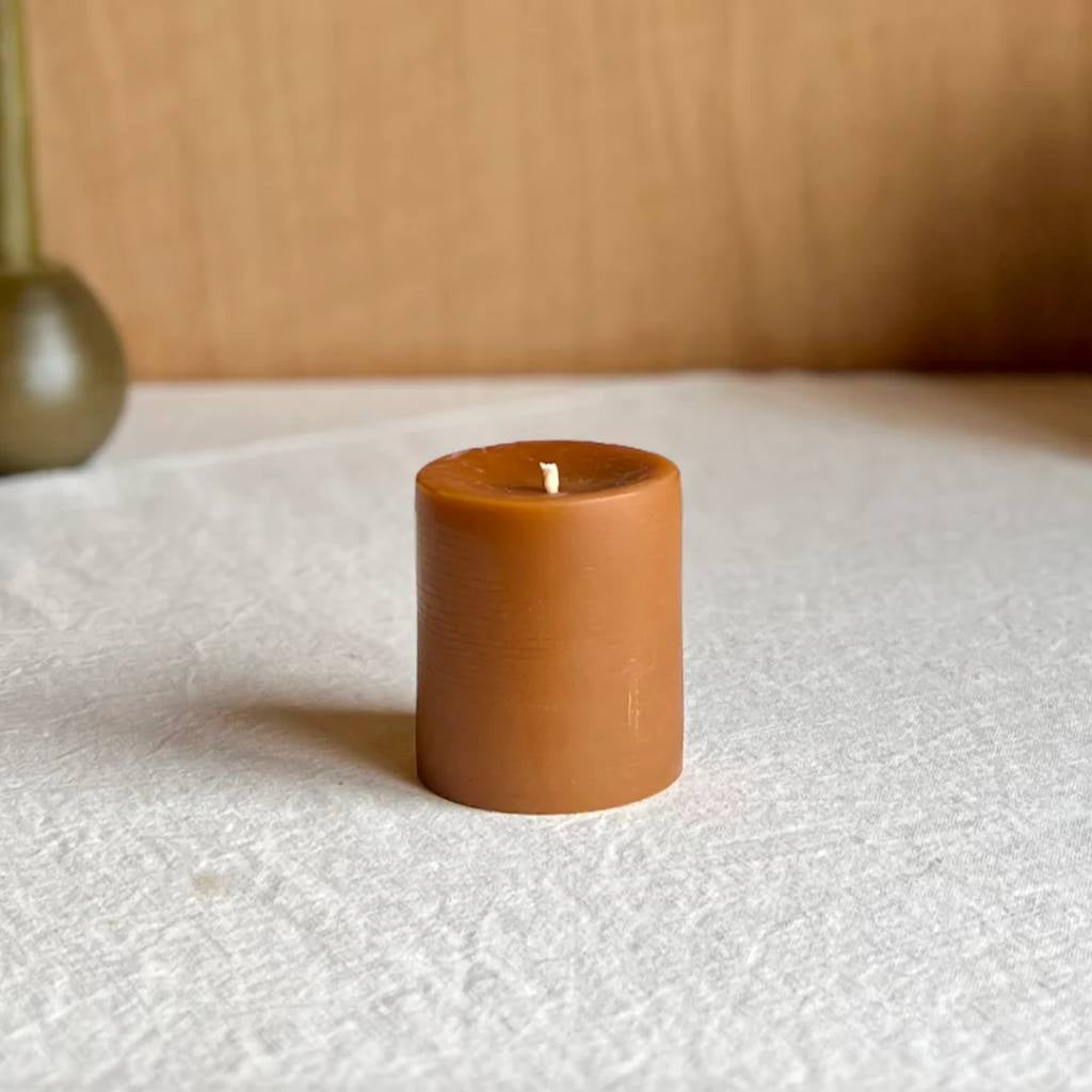 A small amber-colored pillar candle from The Authentic Honey Co., set against a plain background, showcasing its smooth texture and warm hue.