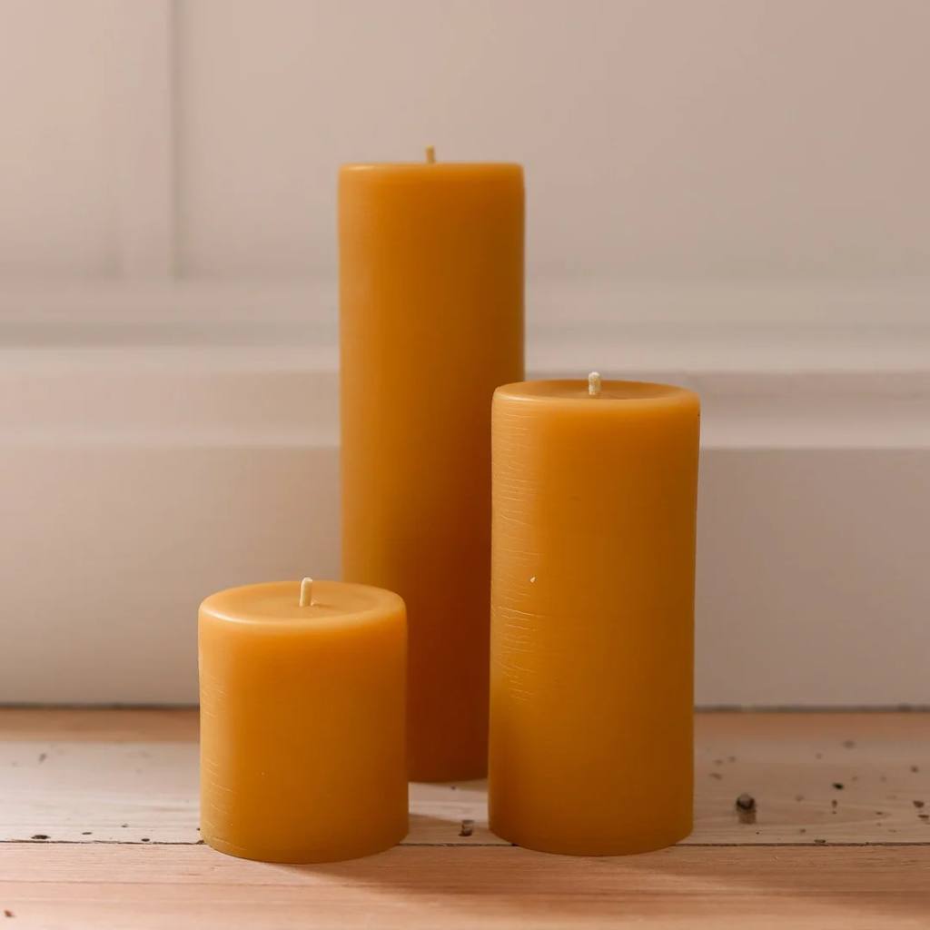 A set of small, natural beeswax pillar candles from The Authentic Honey Co. displayed on a wooden surface. The candles have a smooth, creamy texture and a warm, golden hue, reflecting their natural composition. They are arranged in a simple, elegant manner, highlighting their artisanal craftsmanship and eco-friendly appeal.