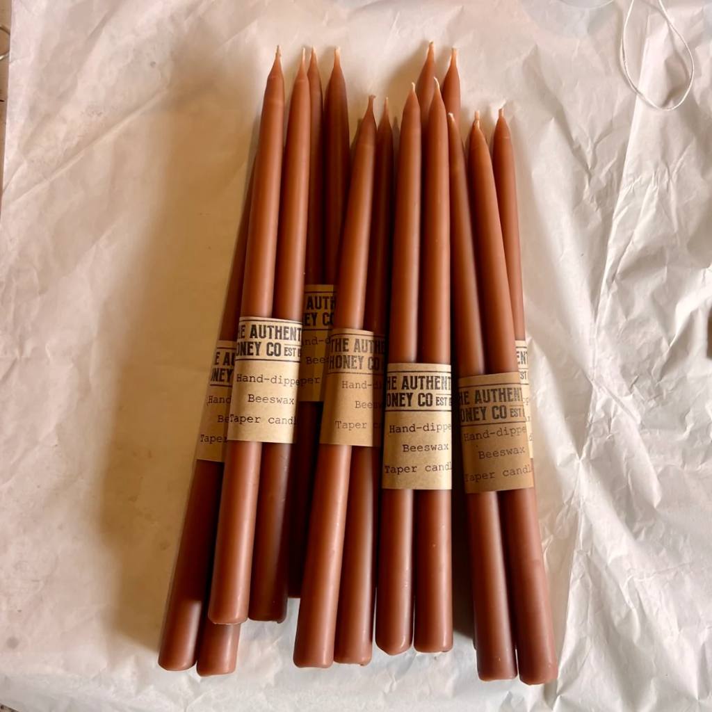 A set of amber-colored taper candles from The Authentic Honey Co. displayed elegantly on a wooden surface. The candles have a smooth, glossy finish and are arranged in a neat row, showcasing their uniform shape and rich, warm hue. The background is softly blurred, highlighting the candles as the focal point of the image.