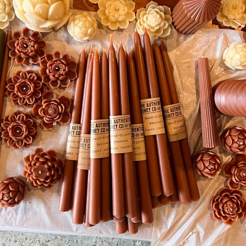 A set of amber-colored taper candles from The Authentic Honey Co. displayed elegantly on a wooden surface. The candles have a smooth, glossy finish and are arranged in a neat row, showcasing their uniform shape and rich, warm hue. The background is softly blurred, highlighting the candles as the focal point of the image.