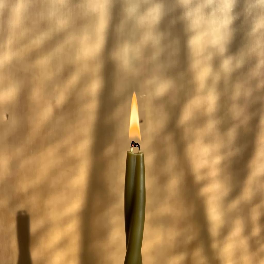 A lit olive-colored twist candle from The Authentic Honey Co. against a neutral background. The candle's unique spiral design is highlighted by the warm glow of the flame, creating a cozy and inviting atmosphere.